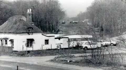 Jack Rose Collection A black and white photo of a house with a caravan and cars parked to the back of it, with construction going on in the background. 