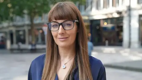 A woman with long brown hair and glasses stands on a Cardiff street, the background is blurred