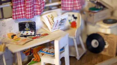 Another dolls house which is more modern with white chair and table, on which sits a miniature set of paints and colouring pens with disney characters coloured in around it. in the background there is a record player with miniature vinyl record discs dotted around.