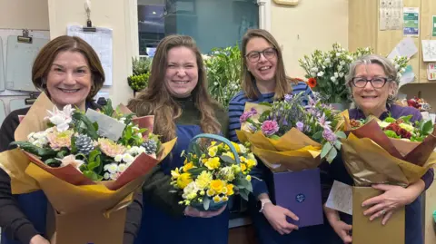 ELLEN KNIGHT/BBC Four women stood side by side - each holding a bouquet of flowers. Jean, on the left, is wearing a dark grey long sleeve top and a navy apron, and is holding a bouquet of pink and white flowers. Natalie, next to her, is wearing a dark green top and a navy apron, holding a blue basket with yellow flowers in. Bea, next to Natalie, is wearing a blue and navy striped top and is holding a bouquet of pink and purple flowers. Catherine, on the right, is wearing a purple long sleeve top and is holding a bouquet of red and green flowers. All the bouquets are wrapped up in brown paper and have gold and purple boxes. 
