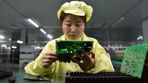 Getty Images A women in a yellow attire holds a green circuit board in a production facility in East China