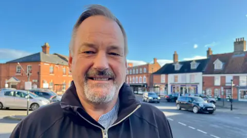 Jo Thewlis/BBC Martin Skoyles has short, grey hair and a small, grey beard. He is wearing a blue jumper with a striped blue shirt underneath.
He is standing in Aylsham market place with parked cars and shops behind him.