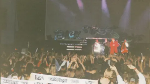 Freedom Leisure A grainy photograph showing the Backstreet Boys on stage with the band name behind them in silver letters. The photo is taken from far back in the crowd, which can be see with their hands in the air and holding up Backstreet Boys scarves