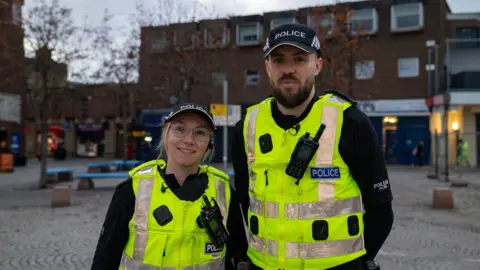 PCs Carolanne Aitken and Daryl Shirkie stand in the middle of a high street - there are shops and businesses behind them. On the left is PC Carolanne Aitken, she is smiling and wearing large glasses. She has blonde hair and is wearing a police Scotland cap and a high vis Police Scotland vest. On the right is PC Daryl Shirkie. He has a brown beard and is wearing a police Scotland cap and a high vis Police Scotland vest.