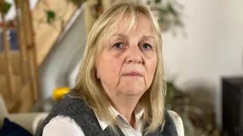 BBC Nicky Jones is looking into the camera.  She is wearing a white shirt and a knitted grey jumper.  She has blonde hair. A TV, plant and staircase are blurred in the background.