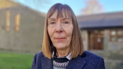 Jill Scott with shoulder length brown hair wearing a purple coat and navy blue, pale blue and pink patterned jumper stands in front of a home blurred out in the background.
