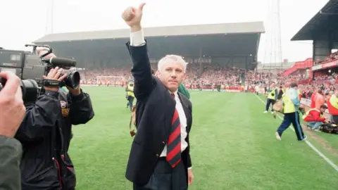 Peter Reid standing in a black suit with a red and black tie. He is stood on the pitch at Roker Park giving a thumbs up to the crowd. 