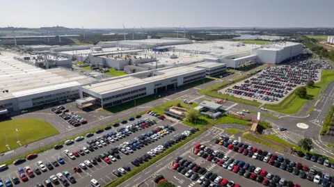 Nissan An aerial view of the the Nissan factory. The large grey and white factory made up of several buildings stretches into the distance. A huge car park is filled with cars.