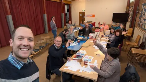 St Thomas Church A man with brown hair smiles in a selfie with a long table in the background where seated people are waving. He is wearing a black jumper with grey stripe and blue checked shirt. The people sitting down are a mixture of men and women. They have stacks of books and paper in front of them. A woman is standing and waving in the background. they are in a hall, with chairs stacked and bulletin boards on the wall. Red curtains are on the left.