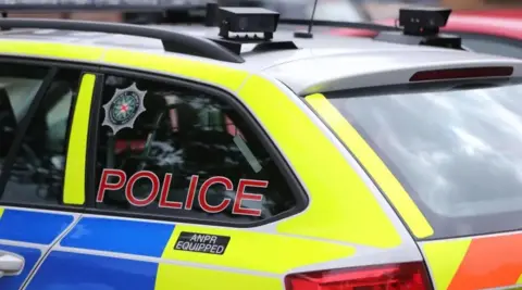A PSNI vehicle, it is blue and yellow and orange. The back left window has a sticker of the PSNI badge. 
