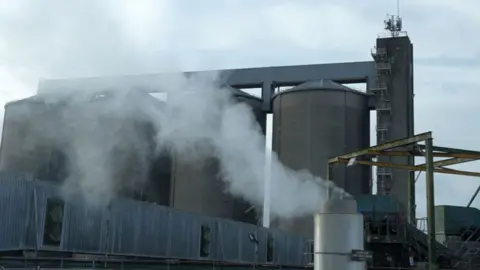 Jamie Niblock/BBC An exterior view of a sugar beet factory. There are four huge silver coloured silos with a low lying shed in front of them. In front of that on the right is a chimney which is blowing smoke or evaporated water. 