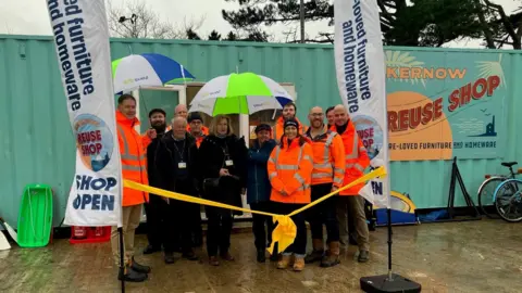 Nine people in orange high-vis coats and three people in black stand outside the entrance to the re-purposed shipping container under two large white, blue and lime green umbrellas. A green plastic sled and the bottom of two fire extinguishers are propped against the building to the left of the group of people.