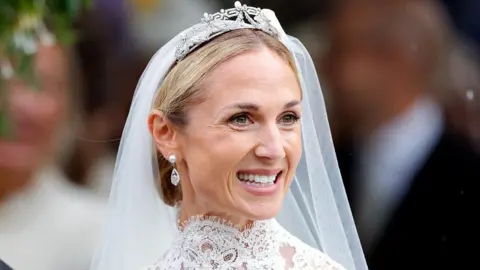 Harriet Sperling, wearing pearl earrings with a silver drop, a veil and a tiara, smiles. She's wearing a high-necked wedding dress