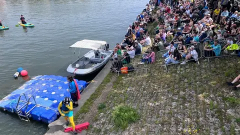 Crowds sat near water with person dressed in yellow climbing out of Bristol Harbourside.