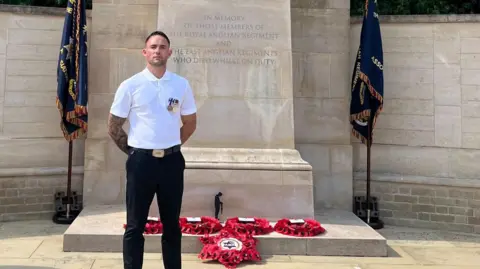 Rhys Thurtell Rhys Thurtell wearing a white shirt and black trousers standing in front of a cenotaph and five read wreaths of poppies 