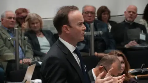John Fairhall/BBC Richard Rout is wearing a black suit and tie with a white shirt. He standing in the council chamber. He is speaking and is animated. 