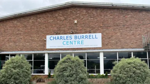 External view of a brick building with 3 large shrubs in front of it. The sign says 'The Charles Burrell Centre. Uniting Community, Learning & Business Under One Roof".