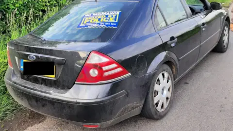 A black car parked near Bristol Airport is seen with a blue and yellow "police aware" sticker on it