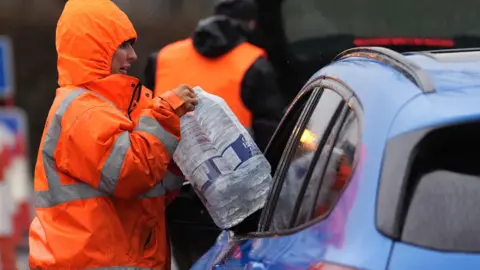 PA Media A woman in a orange high-vis jacket with her hood up, passes a pack of six bottles of water through an open car window.