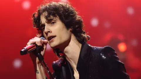 Getty Images Sombr in a shiny dark jacket singing into a microphone on a leaning stand in front of a red background at the 2025 MTV Video Music Awards