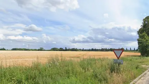Google Maps A landscape image taken of a large open farmer's field from a road. There is a give way sign on the right of the image, which is positioned amongst green fauna. The blue sky is overcast with some clouds. 