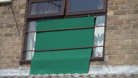 BBC News The photo shows an upstairs window of a house. There is a green sheet which covers the majority of the surface area. It's held there with two brown straps.