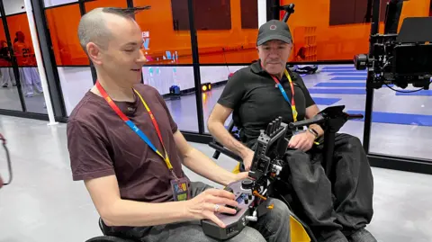 Owen Tooth sat in his wheelchair next to Chris Lynch in the camera system. Tooth is holding Lynch's control panel and monitoring his camera.