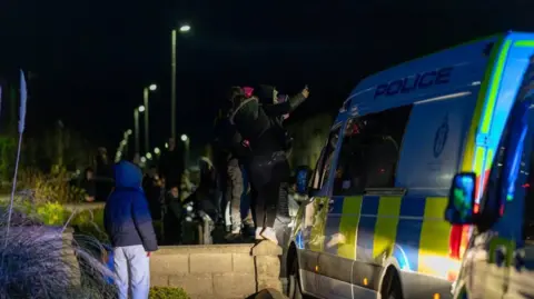 Jasper Images a crowd with hoods up standing on a wall next to a police van