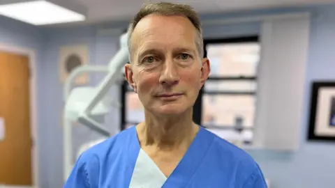 Dentist David McColl, who is wearing bluish  scrubs and is lasting  successful  a dental surgery, looks into the camera