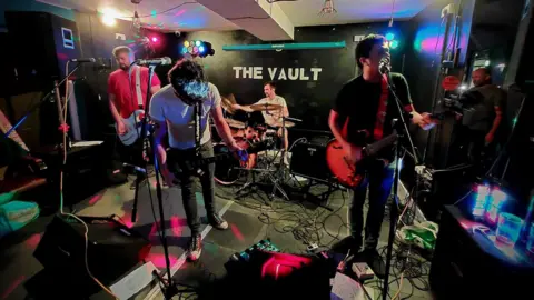 Josh Booth Three men playing guitar and one playing drums perform on a small stage