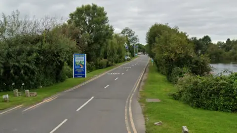 A Google Streetview image of a road lined with trees with a blue sign next to it. It says Welcome to Lakeside on it.