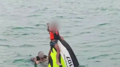 A man waves in the water next to a sunken jet ski which has a young boy sitting on top of it. Just the top triangle of the jet ski is visible above the water. Both the man and boy are wearing life jackets