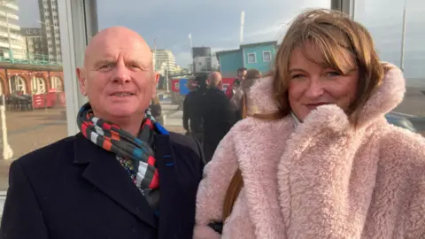 A white man with no hair wearing a stripey scarf looks at the camera and stands next to a white woman with brown hair and a big pink fluffy coat.