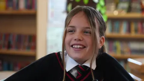 A schoolgirl in a uniform with shelves of books in the background. Her hair is in a ponytail but she has pulled forward two light brand strands either side of her face. She is looking past the camera and smiling.