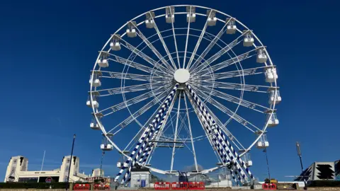 Matt Marvel/BBC A huge white Ferris wheel against a deep blue sky on a seafront. There are red barriers around it.