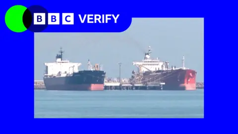 Two cargo ships docked at a port near Iran. Both ships have red bases and are tied to the dock with several ropes. 