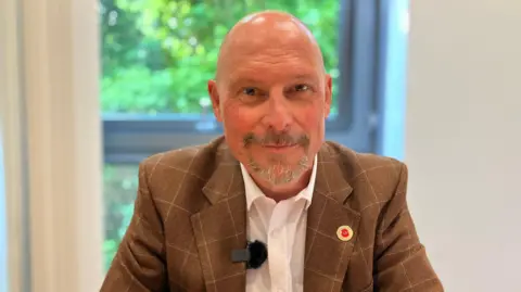 Adam Kent is bald with a goatee and sits behind a desk with a window behind it. He is wearing a brown, checked suit, poppy broach and white shirt.