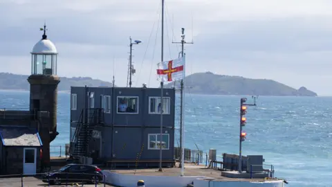 BBC Guernsey flag flies in the wind at the island's harbour entrance. The flag is on a white pole in front of a grey control room. There is sea all around the harbour entrance and the sky appears cloudy.