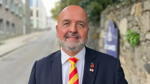 BBC Deputy Jonathan Le Tocq - A bald man with a grey and black beard wearing a suit with a white shirt and red and yellow tie. 
