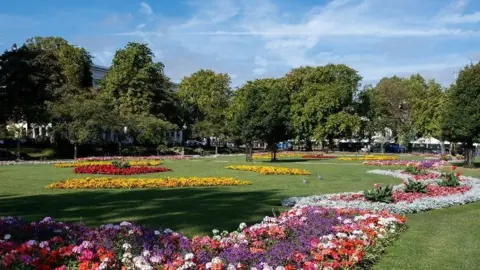 Google Imperial Gardens as seen in the summer. The sky is blue with wispy clouds, and the grass is a lush green. There are lots of bright and colourful flower displays and mature trees dotted around the edge of the park.