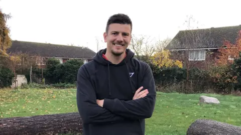 Darren Rozier/BBC A wider view of the man with short dark hair smiles at the camera. He has a small amount of facial hair and is wearing a black hoodie with a red top underneath. He is standing outside and houses can be seen in the distance behind him.