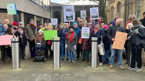 Around 30 protesters pose for a photo outside a council building. Several are holding placards and signs demanding their nature reserve is protected and most are wearing hats and gloves.