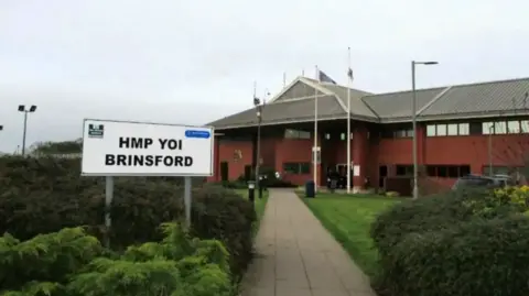 A sign including HMP YOI Brinsford in black letters in the foreground is near bushes. A building, including a grey roof at an angle to the wall for the first and ground floor, is in the background. 