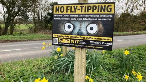 An anti-flytipping sign next to a road with large owl eyes saying that there is surveillance