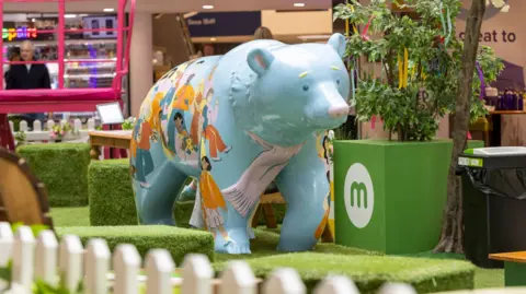 Wild in Art/David Oates Photography A large cartoon-like, light-blue bear sculpture stands on artificial grass inside a shopping centre. Stalls, a white picket fence, green cubes of grass and small trees can also be seen. The bear is decorated with cartoon images of children wearing bright clothes.