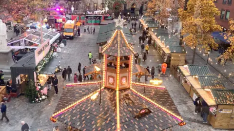 BBC/Phil Cunliffe Picture taken out of the window of a ferris wheel, looking over a market with wooden huts and fairy lights below.