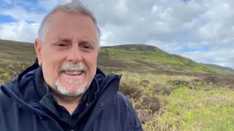 Mike Pratt is a man in his 60s with a grey goatie and moustache and wearing a blue coat and top. There is a hill behind him with grey cloud and moorland shrubbery