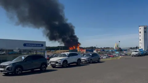 BBC A large fire is tackled at an industrial unit. There are cars parked near the site and orange flames and a large black plume of smoke can be seen.