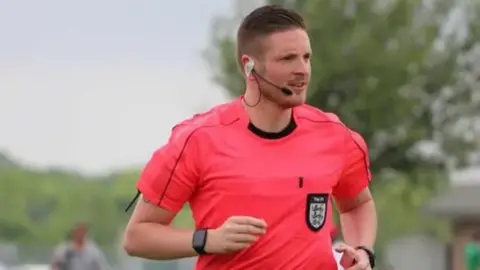 A picture of Ryan wearing a pink referee shirt running. He is wearing a microphone on his face and is wearing a watch on both of his wrists. 