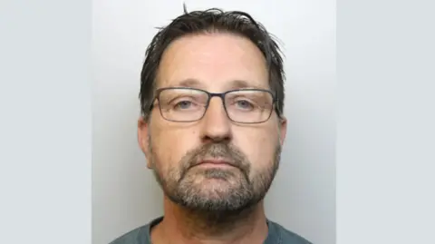 A police custody mugshot of Kevin Thompson, who has a beard and short hair and is wearing glasses.